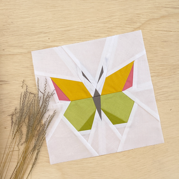 A quilt block, made from paper piecing, with white background fabric and a butterfly in shades of yellow, pink and green.